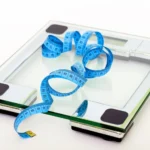 A digital bathroom scale with a blue measuring tape draped across its glass platform on a white background
